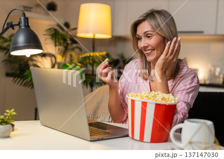 Adult woman with laptop and popcorn watches trailer laughs loud comments funny moments online joyful 137147030