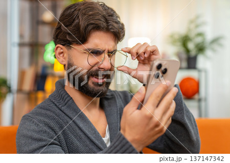 Indian man with eyeglasses squints at smartphone screen tries to read small text poor vision focus 137147342