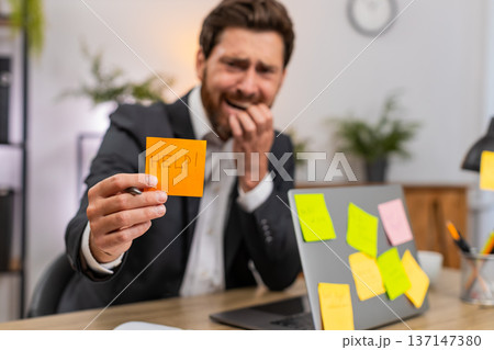 Middle-aged businessman worried by sticky notes on laptop stressed by tasks and overdue plans sighs 137147380