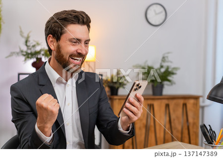Middle-aged businessman texting happy news on smartphone celebrates project win with victory gesture 137147389