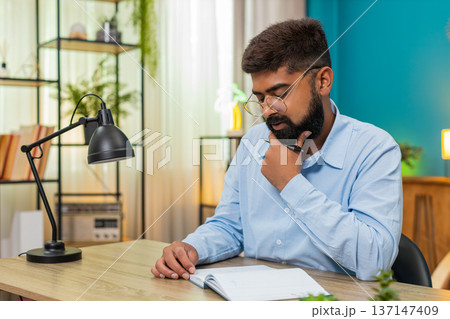 Indian man holds pen gazes notebook deeply thinking planning ideas writing strategy indoors focused 137147409
