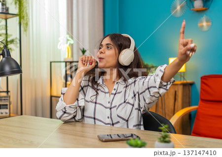 Indian woman sings with headphones using imaginary microphone smiling enjoying favorite song happily Indian woman sings with headphones using imaginary microphone smiling enjoying favorite song happily 137147410