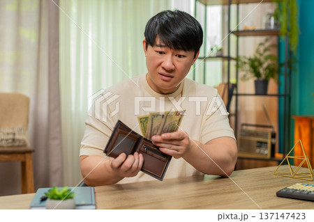 Asian man frowning at nearly empty wallet, sees only coins, slowly shakes head with mild discontent Asian man frowning at nearly empty wallet, sees only coins, slowly shakes head with mild discontent 137147423