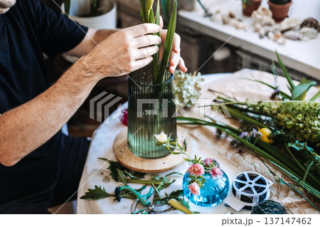 Hands of florist place long green stems into glass vase on table with tools and floral supplies. Independent florist, local business owner, small business startup, craft industry, flower shop studio. 137147462