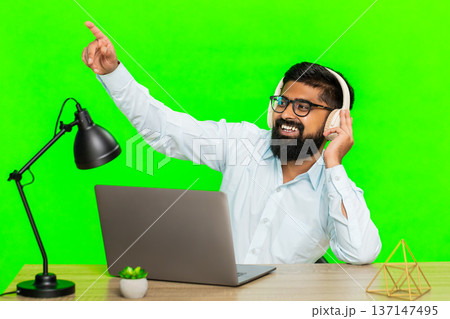 Indian man with wireless headphones working on laptop enjoying music nodding head listening focused 137147495