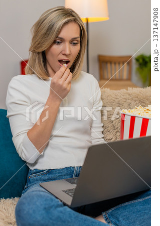 Adult woman stunned by shocking movie moment at home, holding popcorn and staring at laptop screen Adult woman stunned by shocking movie moment at home, holding popcorn and staring at laptop screen 137147908