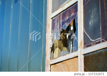 Old window with shattered plastic panel and peeling paint on facade. Neglect, building damage, repair concept and industrial urban background. Old window with shattered plastic panel and peeling paint on facade. Neglect, building damage, repair concept and industrial urban background. 137152071