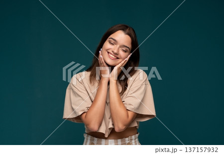 Young woman closing eyes and smiling, hands on face, expressing happiness, joy, and positive emotion for wellbeing 137157932
