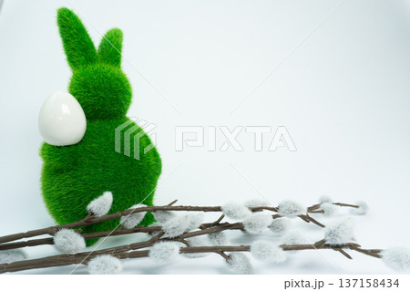 Bright green rabbit holding white egg stands near fluffy branches on white background in spring season 137158434