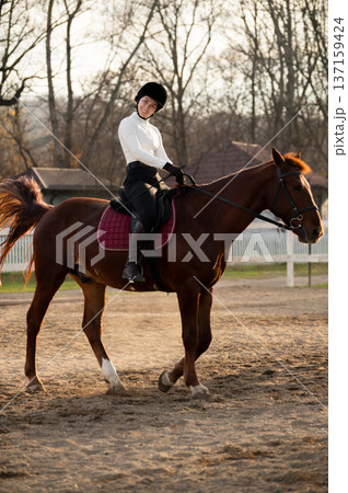 horse rider looks at the camera and smiles in an open arena nearby, sitting in the saddle and wearing a helmet. Horse training and skill development 137159424
