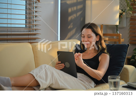 Happy pretty caucasian woman in casual outfit sitting on sofa at home and using digital tablet Happy pretty caucasian woman in casual outfit sitting on sofa at home and using digital tablet 137160313