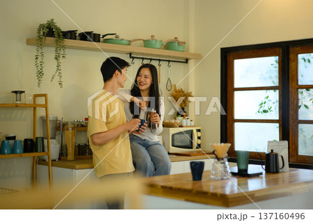 A young couple enjoys a relaxed moment in a warm home kitchen, chatting and smiling while holding coffee, reflecting everyday happiness and modern lifestyle. A young couple enjoys a relaxed moment in a warm home kitchen, chatting and smiling while holding coffee, reflecting everyday happiness and modern lifestyle. 137160496