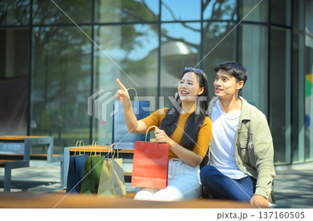 Asian couple sitting outdoors after shopping, smiling and talking together with colorful shopping bags in a modern urban space Asian couple sitting outdoors after shopping, smiling and talking together with colorful shopping bags in a modern urban space 137160505