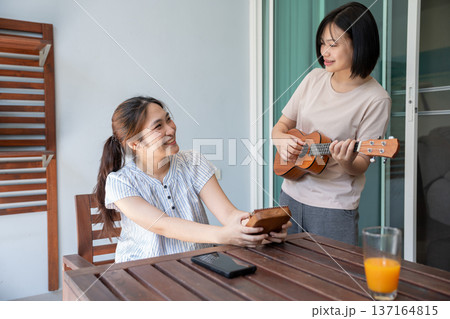 Harmony in the Moment: Two women share a tranquil moment outdoors, one serenading with a ukulele, the other savoring a slice of bread. Harmony in the Moment: Two women share a tranquil moment outdoors, one serenading with a ukulele, the other savoring a slice of bread. 137164815