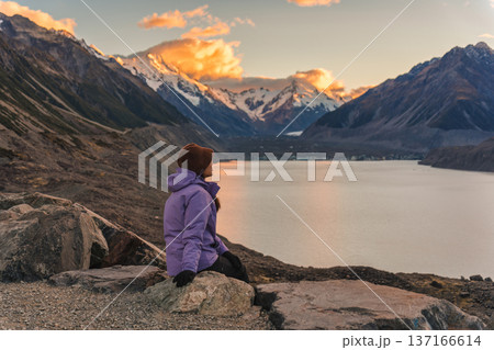 Woman sitting on rock admiring Tasman Lake and snowcapped mountain at sunset in New Zealand 137166614