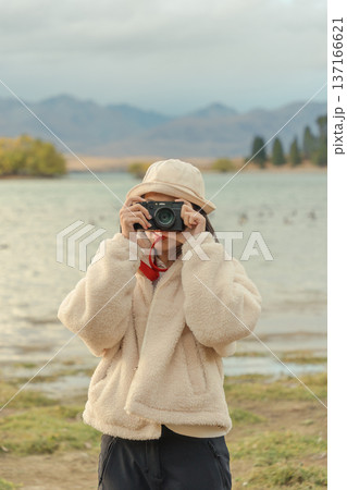 Woman in cozy jacket capturing the moment in autumn nature with retro camera by the lake at New Zealand 137166621