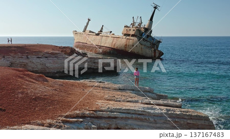 Young girl and others on rocky Paphos coastline watching the stranded Edro III cargo ship against clear blue Mediterranean waters and a bright summer sky, travel and nature clash 137167483