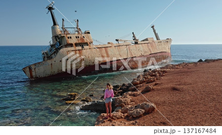 Woman standing on the rocky seashore admiring the iconic rust-covered Edro III Shipwreck grounded on the coast of Paphos, Cyprus, with clear blue Mediterranean sea and sky 137167484