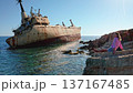 Young woman sitting on a rocky seashore admiring the iconic Edro III shipwreck, a popular tourist landmark near Paphos, under a bright blue sky on a clear day 137167485