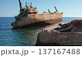 Young Caucasian woman sitting on coastal rocks, looking at the rusty Edro III shipwreck partially submerged in the clear turquoise waters of the Mediterranean Sea near Paphos, Cyprus 137167488