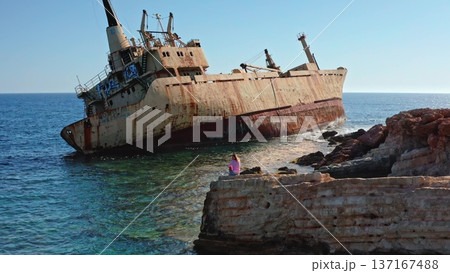 Young Caucasian woman sitting on coastal rocks, looking at the rusty Edro III shipwreck partially submerged in the clear turquoise waters of the Mediterranean Sea near Paphos, Cyprus 137167488