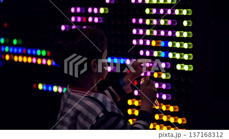 Rear view of a child placing glowing LED pegs into an interactive wall, bright color rows cutting through a dark room. Hands on play builds STEM thinking, focus, and healthy curiosity. 137168312