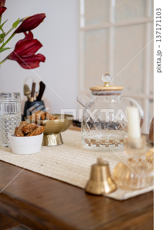 Living space. Home indoor. Transparent teapot glass with tasty wholegrain snack candle bell for sound therapy flower placed on table. Living space. Home indoor. Transparent teapot glass with tasty wholegrain snack candle bell for sound therapy flower placed on table. 137171103