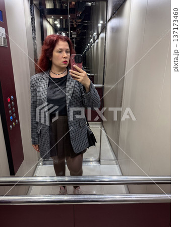 Woman in checked blazer and short brown skirt taking a selfie in elevator. Expression, individuality, and urban confidence captured in a casual moment of everyday life. Woman in checked blazer and short brown skirt taking a selfie in elevator. Expression, individuality, and urban confidence captured in a casual moment of everyday life. 137173460