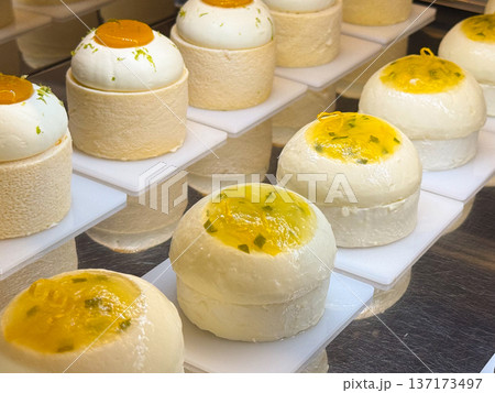 Round mousse cakes topped with mango glaze placed in a pastry display close-up. Dessert presentation symbolizing freshness, softness, and bakery refinement. Round mousse cakes topped with mango glaze placed in a pastry display close-up. Dessert presentation symbolizing freshness, softness, and bakery refinement. 137173497