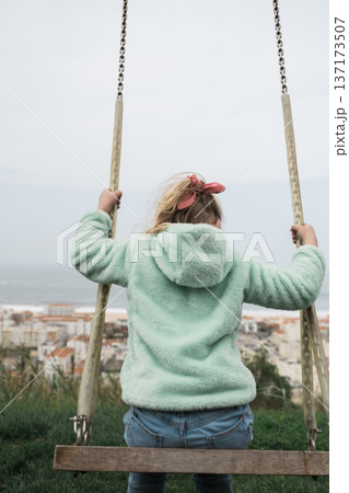 Close up from behind of a young girl with a pink bow in her hair sitting on a swing overlooking a foggy coastal landscape Close up from behind of a young girl with a pink bow in her hair sitting on a swing overlooking a foggy coastal landscape 137173507