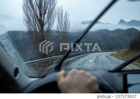 Looking through a rainy windshield at a curving asphalt road leading into foggy hills and snow peaks 137173617