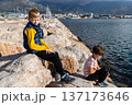 Two boys sitting on rocks by sea at marina, childhood friendship, coastal lifestyle and outdoor leisure 137173646