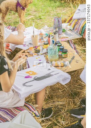 Children decorating white t shirts with acrylic paint and brushes at creative workshop table outdoors, craft session in park. Concept of textile printing, DIY project, art education 137174043