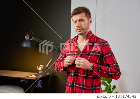 Man standing indoors and buttoning plaid shirt 137174342