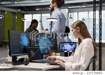 Female computer scientist in office relies on AI to run tasks across multiple devices, monitoring progress on screens. Young tech startup worker doing internship uses neural networks to develop AI Female computer scientist in office relies on AI to run tasks across multiple devices, monitoring progress on screens. Young tech startup worker doing internship uses neural networks to develop AI 137176602