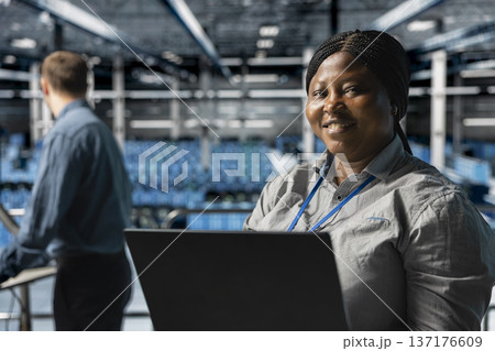 Close up of smiling worker in server farm next to colleague upgrading hardware for increased storage capacity. Jolly african american data center admin and coworker doing maintenance Close up of smiling worker in server farm next to colleague upgrading hardware for increased storage capacity. Jolly african american data center admin and coworker doing maintenance 137176609