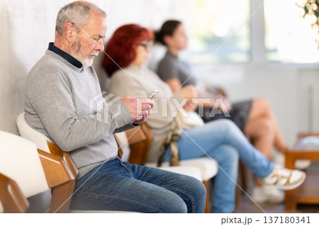 Elderly man sitting in clinic waiting area scrolling mobile phone Elderly man sitting in clinic waiting area scrolling mobile phone 137180341