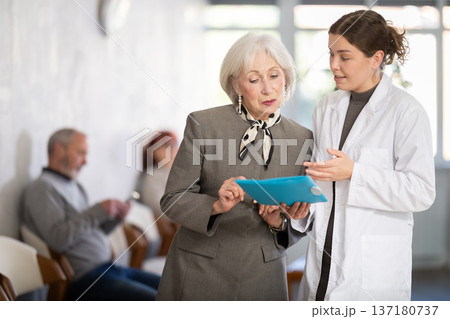 Positive polite nurse or doctor talking to aged woman patient in reception area in background queue of people 137180737