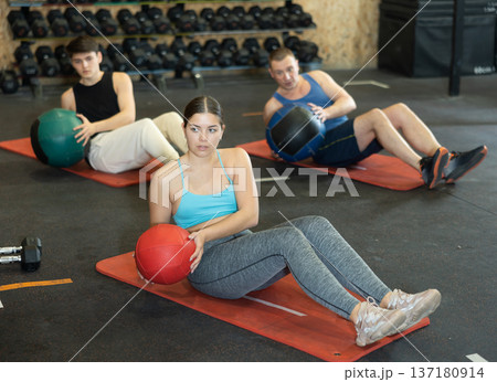 Motivated young woman practicing exercises with medicine ball near other people in gym Motivated young woman practicing exercises with medicine ball near other people in gym 137180914