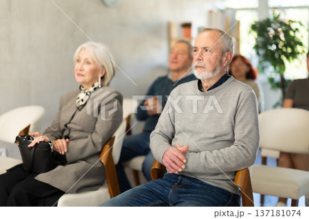 Focused elderly man wearing ordinary clothes sitting on chair with group of other students of different ages listening speech in light conference room at seminar Focused elderly man wearing ordinary clothes sitting on chair with group of other students of different ages listening speech in light conference room at seminar 137181074