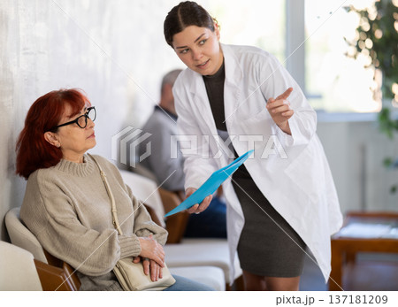 Polite young nurse or doctor talking to mature woman patient standing in waiting room of medical clinic, with her back to visitors sitting in chairs 137181209