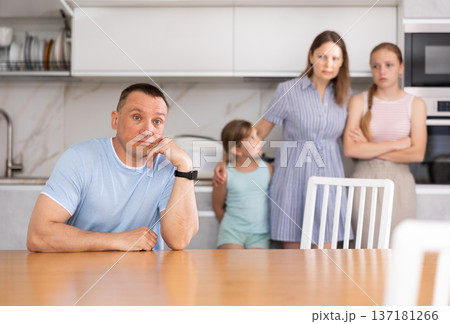 Upset father is sitting at the table, in background is a sad wife with small children. Family quarrel in kitchen Upset father is sitting at the table, in background is a sad wife with small children. Family quarrel in kitchen 137181266