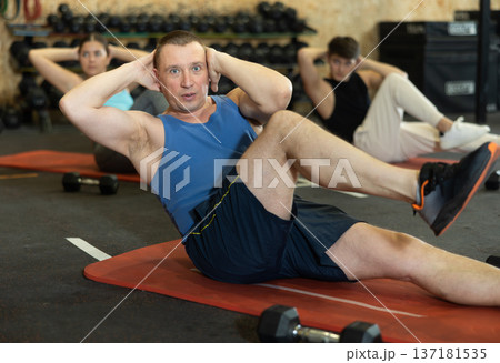 Dedicated man practicing knee raises near other people in gym Dedicated man practicing knee raises near other people in gym 137181535