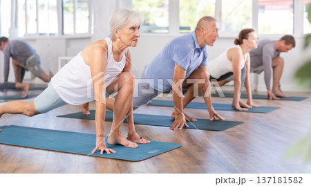 Mature female participant working out practice warrior pose, virabhadrasana with group Mature female participant working out practice warrior pose, virabhadrasana with group 137181582