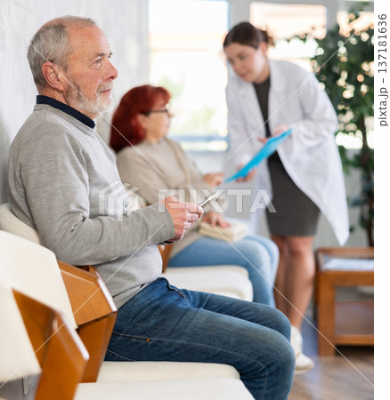 Elderly man sitting in clinic queue while nurse talks to other patients 137181636