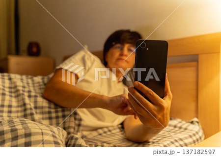 Mature sleepy woman lying in bed browsing social media on smartphone at night, insomnia, internet addiction 137182597