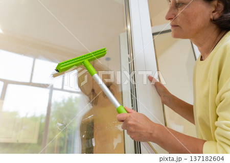 Mature female cleaning windows with green squeegee at home. Cleaning and professional house cleaning, small business 137182604