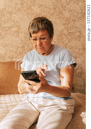 An elderly woman wearing glasses was intently immersed in her smartphone while sitting on a sofa in a sun-drenched. Digital Phone Addiction in Older Women, Screen Time Overload, and Modern Technology An elderly woman wearing glasses was intently immersed in her smartphone while sitting on a sofa in a sun-drenched. Digital Phone Addiction in Older Women, Screen Time Overload, and Modern Technology 137182621