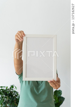 Adult holding blank white frame in green shirt with green plant in background. Modern minimalist design, empty template for text 137182627
