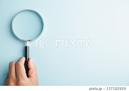 A hand holding a magnifying glass on a light blue background. A hand holding a magnifying glass on a light blue background. 137182929